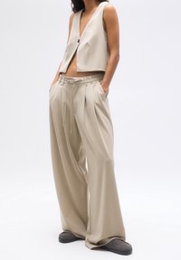 Gilet beige raccourci avec un seul bouton noir, associé à un pantalon ample plissé avec une taille à cordon, fabriqué dans un tissu texturé.