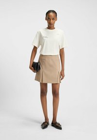 Hvid kortærmet t-shirt med tekst, beige plisseret miniskørt med striber, og sorte spidse loafers, bærende en sort håndtaske.