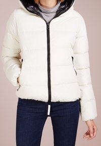 Personne portant une doudoune blanche avec une fermeture éclair et des poches noires, un col roulé gris et un jean bleu marine, se tenant devant un fond clair.