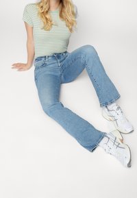 Femme blonde portant un t-shirt rayé vert, un jean bleu clair et des baskets blanches, assise par terre contre un fond blanc.
