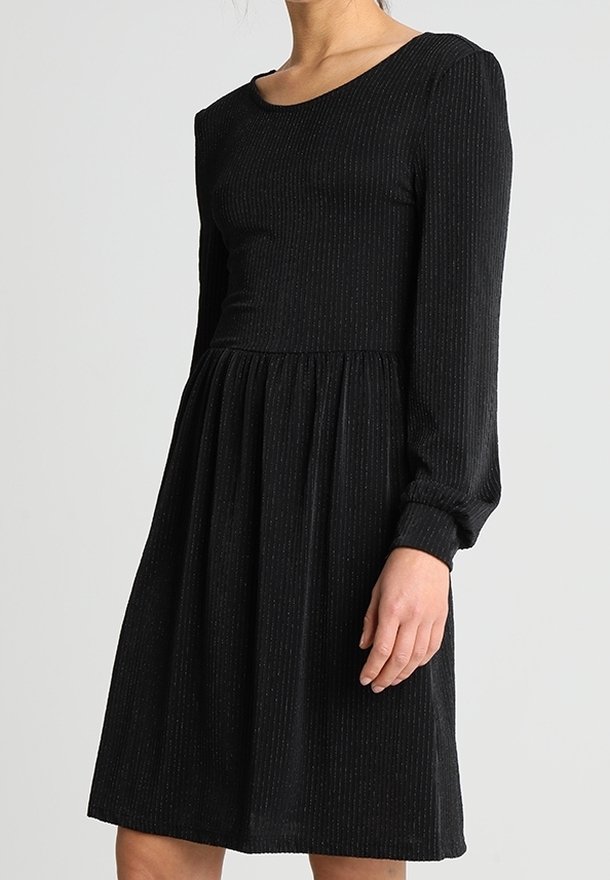 Femme portant une robe noire côtelée à manches longues avec un corsage ajusté et une jupe froncée, debout devant un fond clair uni.
