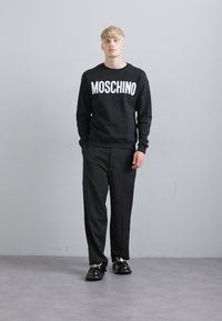 Blond man som står mot en grå vägg, iförd en svart Moschino-tröja, svarta byxor och svarta sneakers, med handen i fickan.