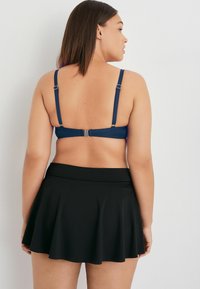 Navyblauer BikinOberteil mit verstellbaren Trägern, kombiniert mit einem schwarzen, ausgestellt geschnittenen Baderock. Glatte Textur, schlichtes Design und sichtbare, sichere Verschlüsse.