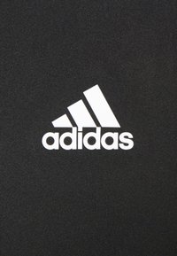 Un tissu noir arbore un logo Adidas blanc, composé de trois bandes diagonales et du nom de la marque en minuscules. Texture lisse et athlétique.