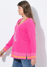 Top lungo a maniche con scollo a V di colore rosa, con grafiche floreali multicolori e dettagli gialli sulle maniche e sull'orlo. Le fessure laterali migliorano la vestibilità.