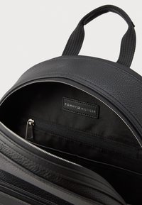 Vnútro čierneho koženého batohu so zipsovým vreckom a značkovou etikettou. Hladká textura s pevnou konštrukciou a látkovými popruhmi.