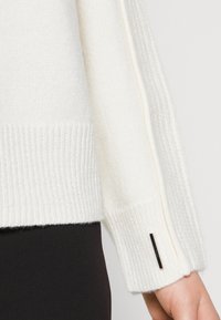 Vit stickad tröja med ribbat mönster, med sidosträck och en svart rektangulär etikett på ärmslutet.