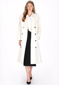 Witte dubbelgeknoopte trenchcoat met knoopdetails, kraag en lange mouwen, gedragen over een zwarte jurk en gecombineerd met zwarte hakken.
