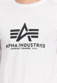 Hvit langermet T-skjorte med et svart grafisk logo som har en stilisert "A" og tekst: "ALPHA INDUSTRIES, KNOXVILLE, TENNESSEE, U.S.A."