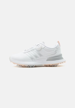 Zapato de golf blanco con rayas plateadas, cierre ajustable de dial y suaves púas en la suela color melocotón sobre un fondo neutro.