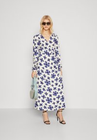 Robe portefeuille fleurie en crème avec des motifs de fleurs bleues, manches longues, décolleté en V et une silhouette fluide. Associée à un sac à main bleu et des talons noirs.