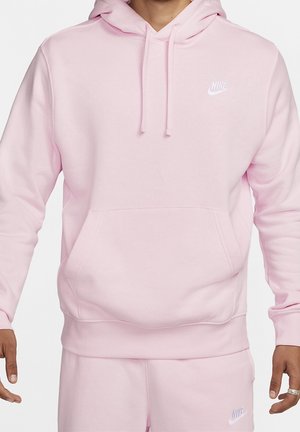 Homme portant un hoodie Nike rose clair et un jogging assorti avec des logos Nike blancs sur la poitrine et la cuisse, mains relâchées le long du corps.