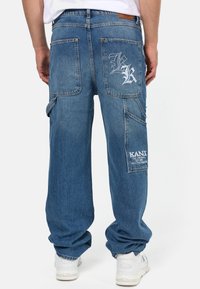 Huppuja siniset denim-farkut, joissa on useita taskuja, brodeerattu suunnittelu ja logopatchi vasemmassa lahkeessa. Vahvistettu ompelu on näkyvissä.