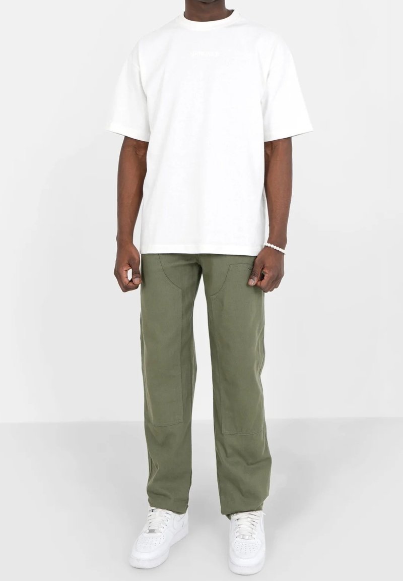 Homme debout portant un T-shirt blanc à manches courtes, un pantalon vert olive avec des renforts aux genoux, des baskets blanches et un bracelet de perles au poignet droit.