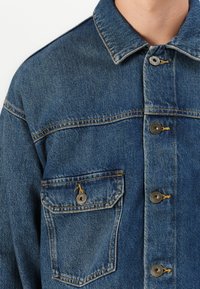 Close-up of blue denim jacket front with metal buttons and a single buttoned chest pocket on a person.