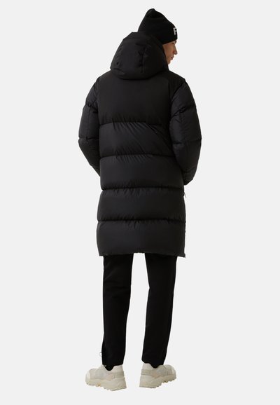 Mann, der mit dem Rücken zu sehen ist, trägt eine schwarze Daunenjacke mit Kapuze, eine schwarze Mütze, schwarze Hosen und weiße klobige Sneakers vor einem einfarbigen Hintergrund.
