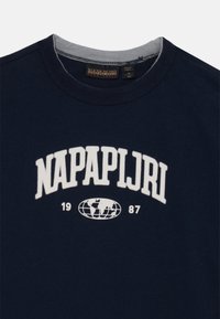 Tamsiai mėlyna medvilninė marškinėliai su apvalia iškirpte, su balta siuvinėta užrašu "NAPAPIJRI" ir žemėlapiu, o žemiau – metai "1987".