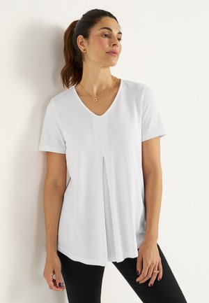 White short-sleeved top with a V-neck, featuring a loose fit and a pleat detail at the center front. Worn with black fitted pants.
