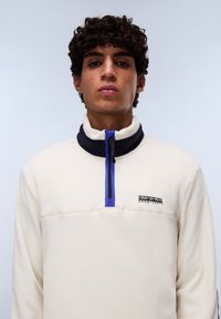 Cream fleece pullover with a high navy collar, purple zip accent, 1/4 zip design, and a small black logo on the chest. Smooth texture.