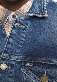 Džínová bunda světlé modré barvy s klasickým límcem, kontrastním prošíváním a malou nášivkou s logem. Vnitřní vrstva odhaluje vzor kárované košile.