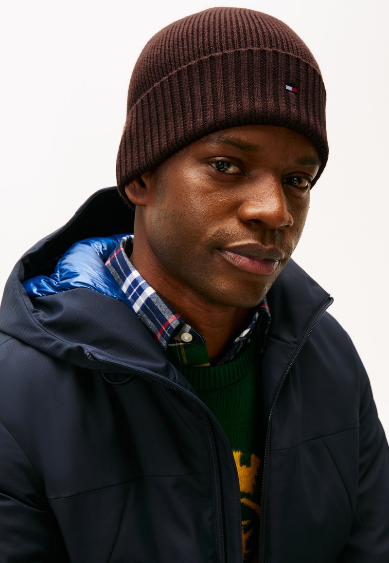 Bruine geribbelde gebreide beanie, marineblauwe jas met een blauwe puffy voering, gelaagd met een groene trui en een geruite blouse, die meerdere texturen laat zien.