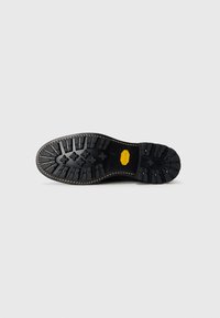 Suela de zapato de goma negra con una superficie texturizada, que presenta un patrón en zigzag y un detalle amarillo, con costuras reforzadas en el borde.