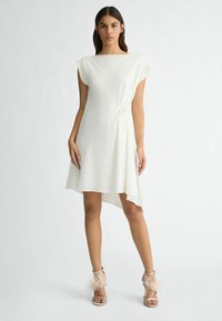 Abito bianco con un orlo asimmetrico, maniche corte a campana e un dettaglio decorativo a cerchio in vita. Abbinato a scarpe con tacco alte con finiture in piuma.