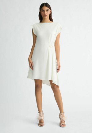 Rochie albă cu o linie asimetrică a tivului, mânecă scurtă tip cap și un detaliu decorativ cu buclă la talie. Purtată cu pantofi cu toc înalt care au accente din pene.