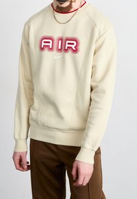 Sudadera beige con un gráfico rojo "AIR", cuello redondo, puños y dobladillo acanalados. Presenta un pequeño logotipo blanco de Nike encima del gráfico.