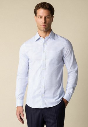 Man met een lichtblauw geruit overhemd en donker geruite broek, staand met één hand in de zak.