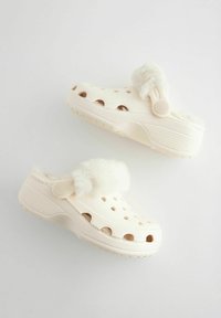 Sabots beige clair avec une douce bordure en fausse fourrure. Fabriqués en plastique moulé, dotés de trous de ventilation et d'une sangle arrière pour un maintien sécurisé.