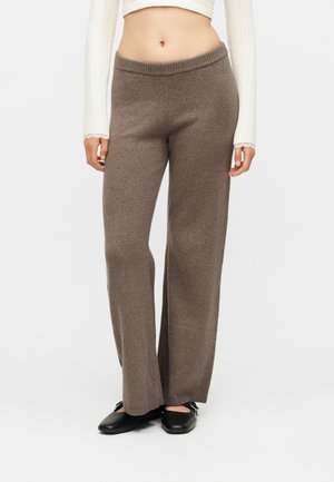 Vrouw die bruine, brede brei-broek draagt, een witte cropped top met lange mouwen en zwarte ballerina's, staand tegen een effen achtergrond.