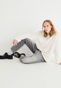 Lett, teksturert hvit strikkegenser, grå jeans og svarte sko med spraglete mønster. Modell sitter med ett ben bøyd, nøytral bakgrunn.