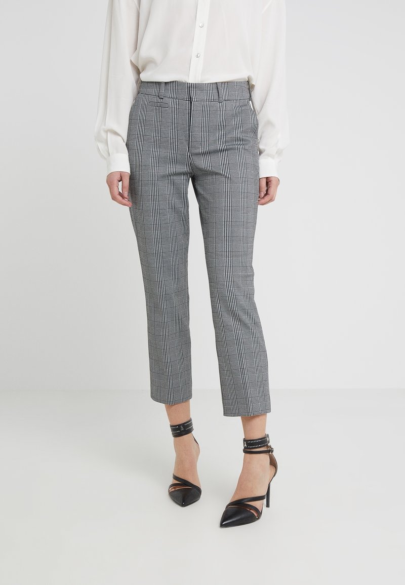 Femme portant un pantalon court à carreaux noir et blanc, un chemisier blanc, et des talons hauts noirs à bride à la cheville sur un fond uni.