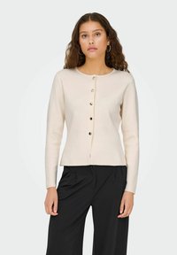 Cardigan beige clair à manches longues, col rond et six boutons dorés sur le devant. Tissu doux et coupe décontractée.