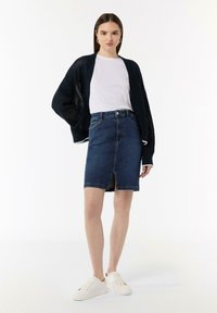 Denimrock mit einem Schlitz auf der Vorderseite, kombiniert mit einem weißen T-Shirt und einer navyblauen Strickjacke. Das Model trägt weiße Sneakers.