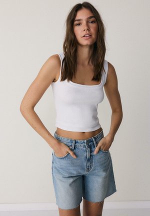 Jeune femme aux longs cheveux bruns portant un top court blanc sans manches et un short en jean bleu clair, mains dans les poches, fond neutre.