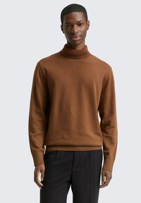 Pull à col roulé en maille marron avec des poignets et un ourlet côtelés, présentant une texture lisse. Le modèle porte un pantalon noir.