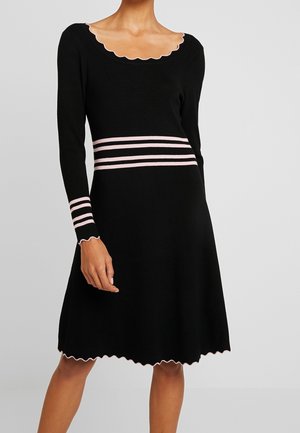 Kobieta ubrana w czarną sukienkę z długim rękawem, z wykończeniami w kształcie falbanek oraz różowymi paskami na wysokości talii i mankietach, stojąca na jednolitym jasnym tle.