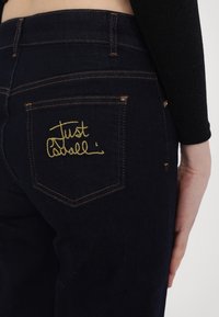 Tmavomodré denimo džiny so priliehavým strihom. Obsahujú zadnú kapsu so zlatou vyšívanou textom "Just Cabell" a kontrastným oranžovým prešívaním.