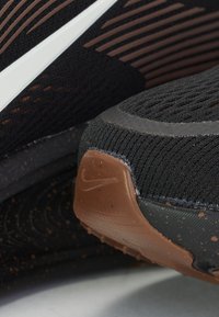 Black athletic shoe featuring a textured knit upper with brown accents, a flexible sole with a distinctive tread pattern, and a small Nike logo.