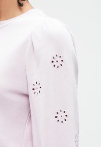 Lichtpaarse katoenen top met pofmouwen, versierd met bloemvormige uitsparingen en fijne afwerkingen aan de randen.