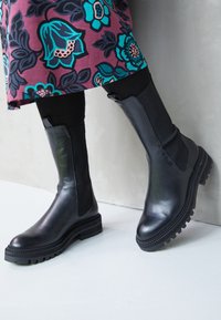 Mustat nahka nilkkabootsit, joissa on joustavat sivupaneelit ja tekstuuriset kumipohjat. Ominaisuuksiin kuuluu sileä pinta ja ommeltu yksityiskohtia reunojen ympärillä.