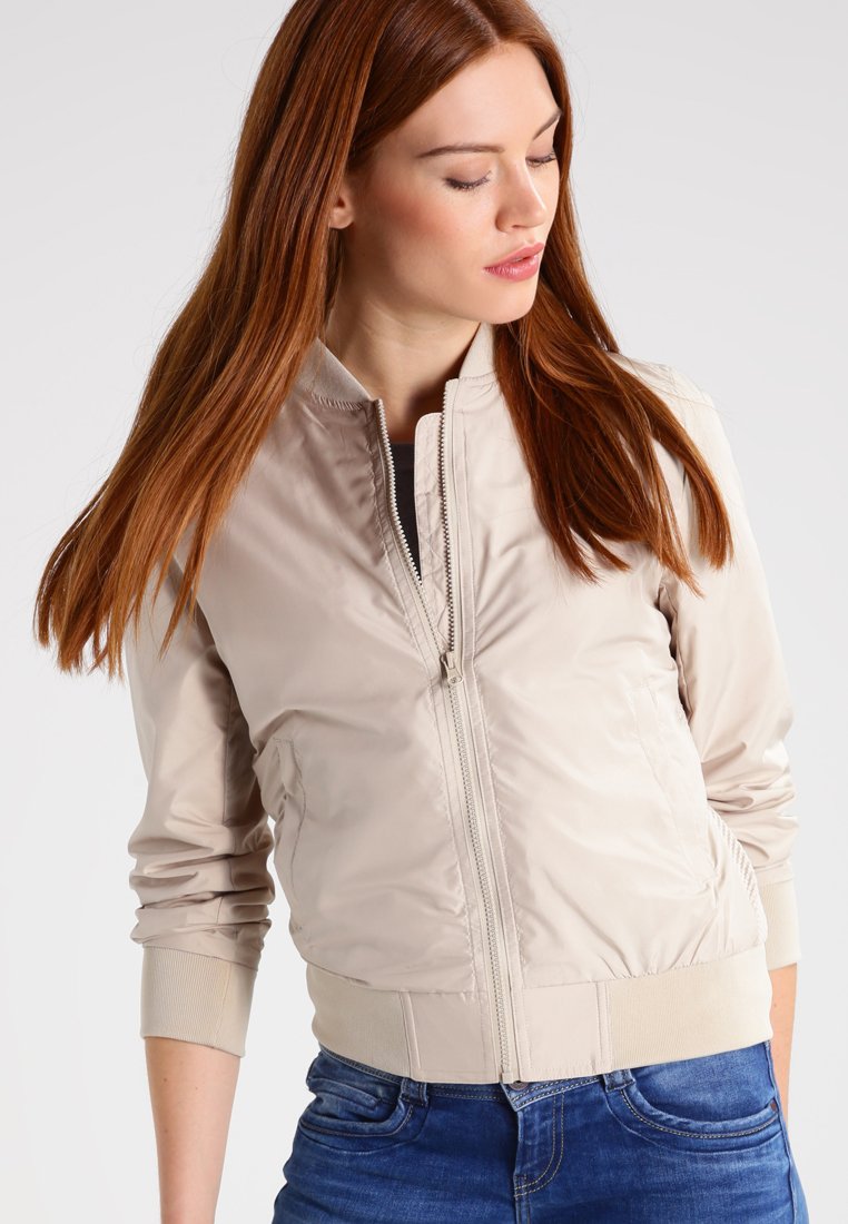 Lichtbeige bomberjack met een gladde, glanzende textuur, ritssluiting, geribbelde manchetten en een aansluitende taille, gestyled met een blauwe spijkerbroek.