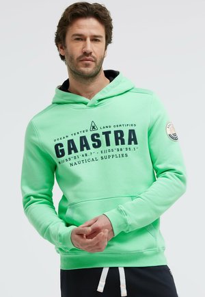 Mann trägt einen hellgrünen Gaastra-Hoodie mit nautischen Koordinaten und Logo, steht vor einfarbig hellem Hintergrund.