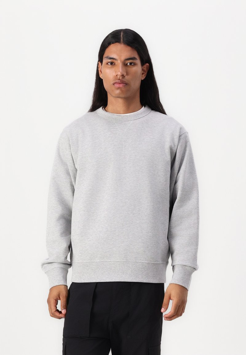 Felpa grigio chiaro in misto cotone, con scollatura a girocollo, maniche lunghe, polsini a coste e vestibilità comoda. Indossata con pantaloni cargo scuri.