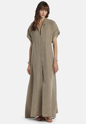 Žena v dlhej voľnej béžovej ľanovej šaty s krátkymi rukávmi, hlbokým výstrihom do V a sandálmi, stojaca pred jednoliatym svetlým pozadím.