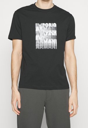 Homme portant un t-shirt noir avec un design textuel blanc déformé "Emporio Armani", associé à un pantalon gris foncé, debout devant un fond uni.
