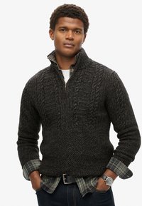 Schwarzer Strickpullover mit halbem Reißverschlusskragen, über einem karierten Hemd getragen. Strukturierte Webart, eng anliegende Bündchen und kombiniert mit Jeans. Silberne Armbanduhr als Akzent.