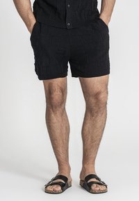 Shorts neri a maglia con un motivo testurizzato, dotati di una vita elasticizzata e tasche. Abbinati a sandali neri a scivolo su piedi nudi.
